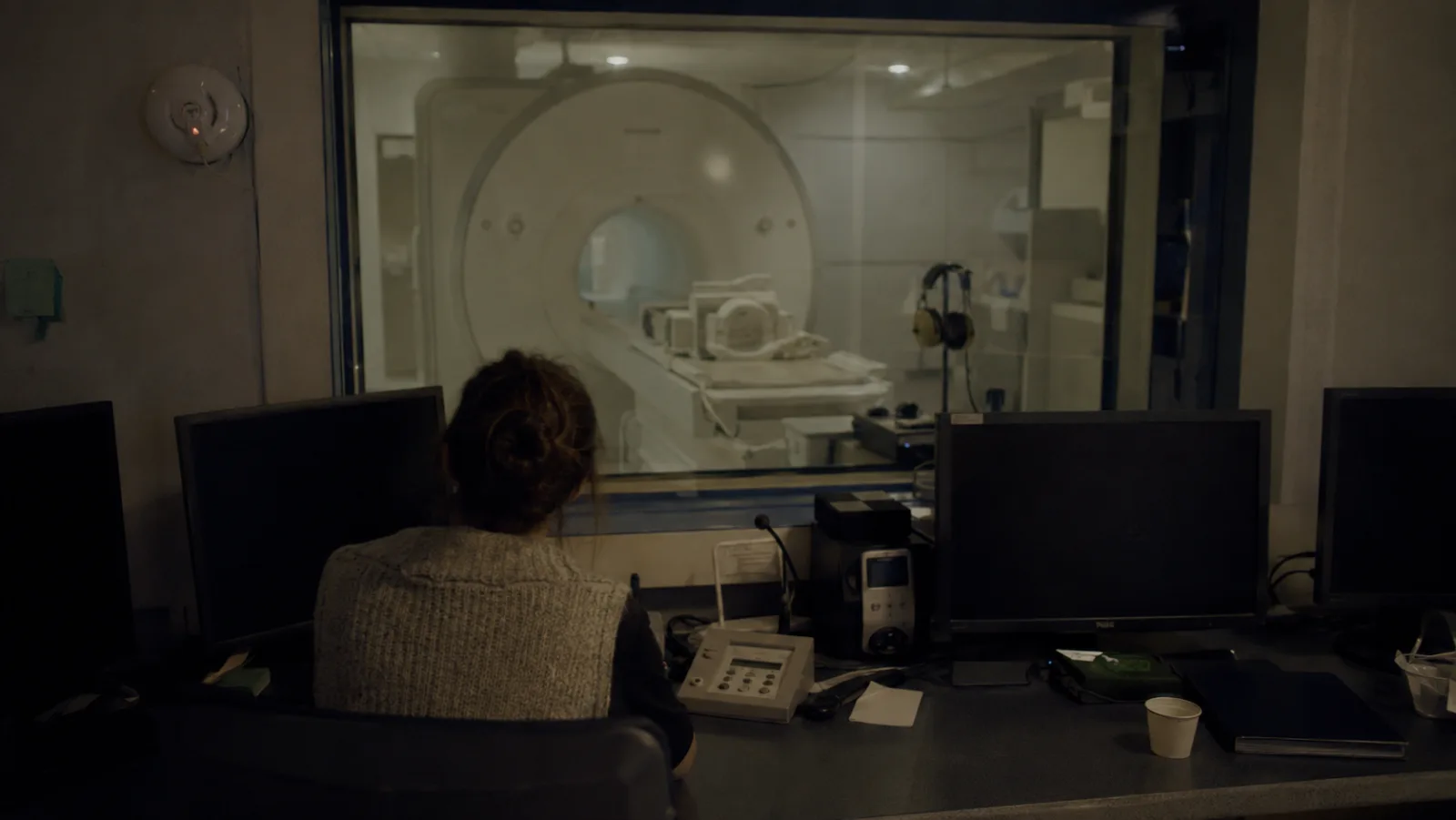 Une technicienne, de dos, observe un appareil d’IRM à travers la vitre d’une salle de contrôle.