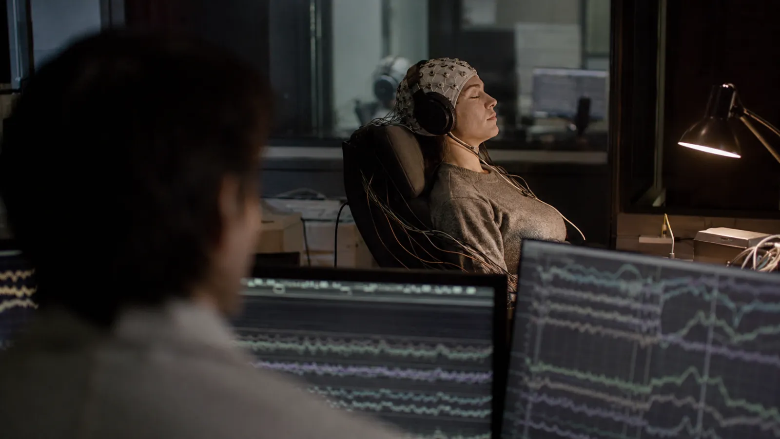 Dans un laboratoire du sommeil, une participante allongée porte un bonnet EEG et un casque audio, yeux fermés ; au premier plan, un chercheur suit les tracés cérébraux sur des écrans.